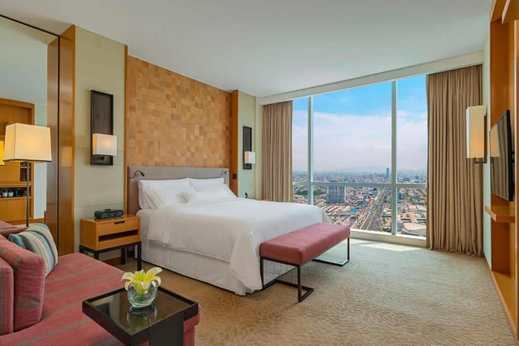 Quarto de hotel com uma cama grande, banco, sofá, vista da cidade através de janelas do chão ao teto e decoração em cores neutras.