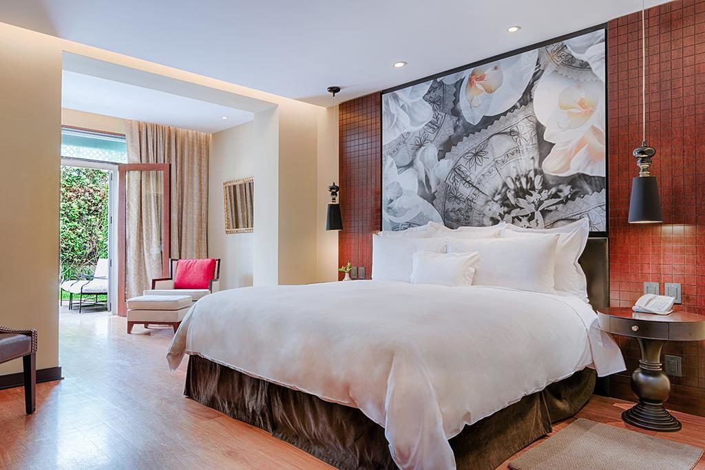 Quarto de casal no andar térreo do Country Club Lima Hotel, um dos hotéis em Lima mais indicados. Um quarto de hotel moderno com uma cama grande, obras de arte florais acima da cabeceira da cama e uma poltrona de lounge com portas de vidro abertas.