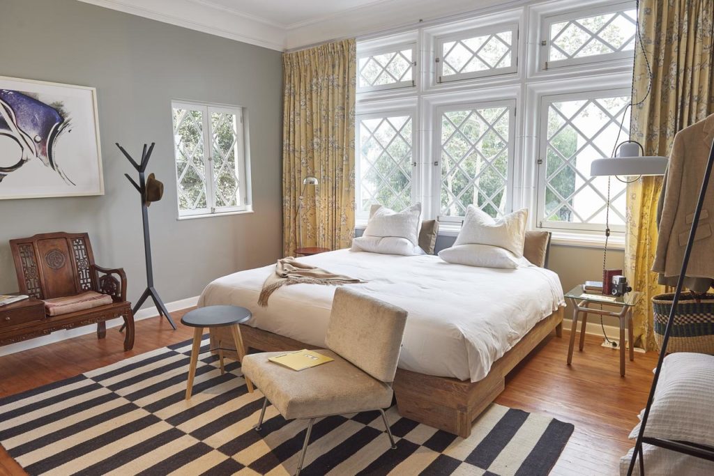 Quarto no Atemporal, um dos hoteis em Lima, com decoração retrô com cama ampla, poltronas, cabideiro, cortinas amarelas e tapete listrado.