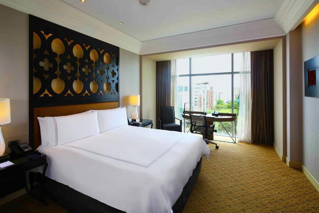 Cama de casal num dos quartos do Hilton Lima Miraflores, um dos hotéis em Lima. Quarto de hotel moderno com uma cama grande, escrivaninha, cadeira, luminárias, cabeceira estampada e uma janela com vista para prédios e árvores.
