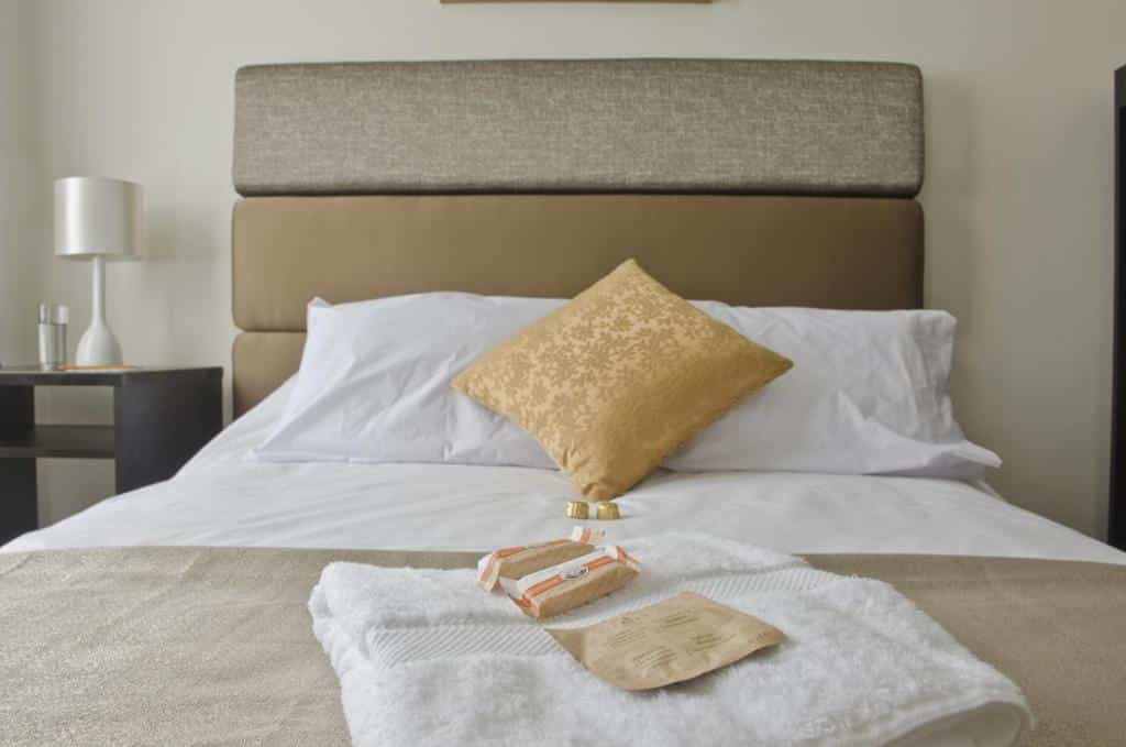 Uma cama bem arrumada com lençóis brancos, um travesseiro dourado e toalhas dobradas com produtos de higiene pessoal colocados em cima.