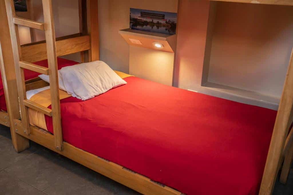 Cama de beliche do Pariwana Hostel, uma das alternativas aos hotéis em Lima. Um beliche inferior bem arrumado com um cobertor vermelho e um travesseiro branco, ao lado de uma escada de madeira e uma luminária de parede.