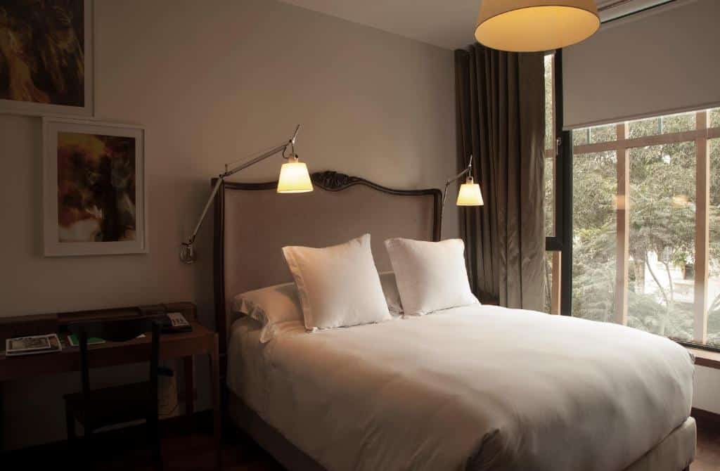 Quarto do Hotel B, um dos hotéis em Lima. Uma cama bem arrumada com roupa de cama branca, dois travesseiros, luminárias de parede, uma escrivaninha e uma grande janela com cortinas em um quarto de hotel.