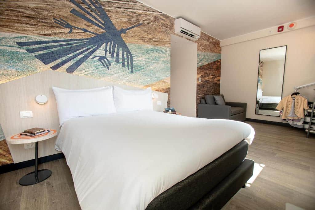 Quarto do ibis Styles Lima Benavides Miraflores, um dos hotéis em Lima. Um quarto de hotel moderno com uma cama grande, arte na parede, uma mesa pequena, um sofá e um espelho que reflete um cabide.