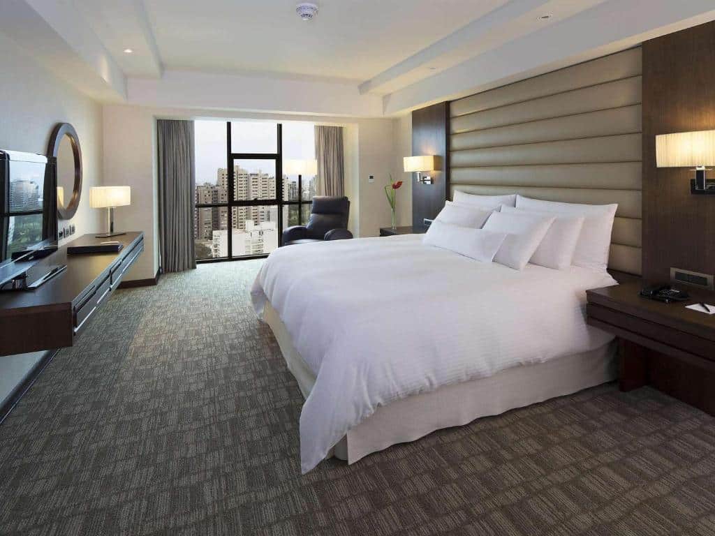Quarto de um dos hotéis em Lima, o Swissotel. moderno com uma cama grande, TV, poltrona e vista da cidade através de janelas do chão ao teto.