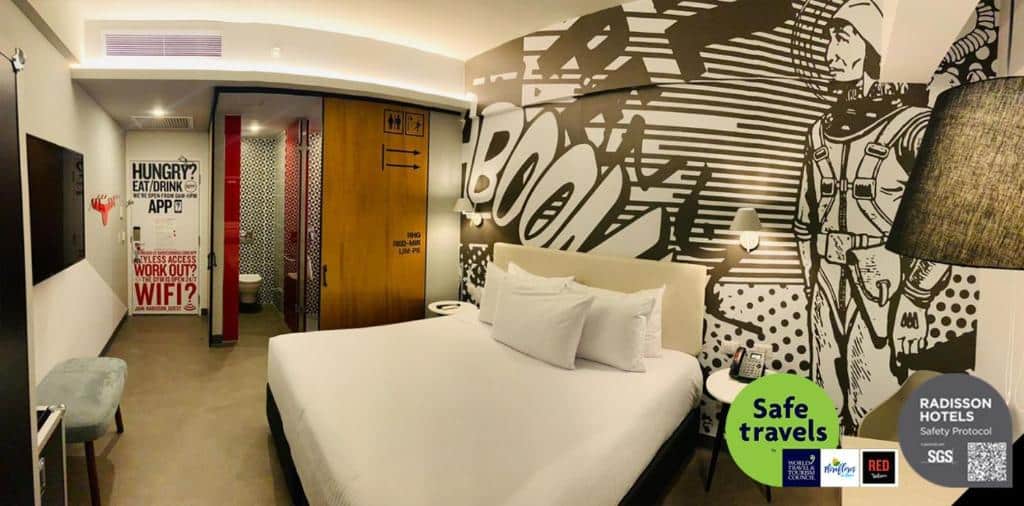 Quarto do Radisson Red Miraflores, um dos hotéis em Lima. Um quarto de hotel moderno com uma cama grande, arte na parede com tema de quadrinhos e logotipos de certificação de segurança no canto.