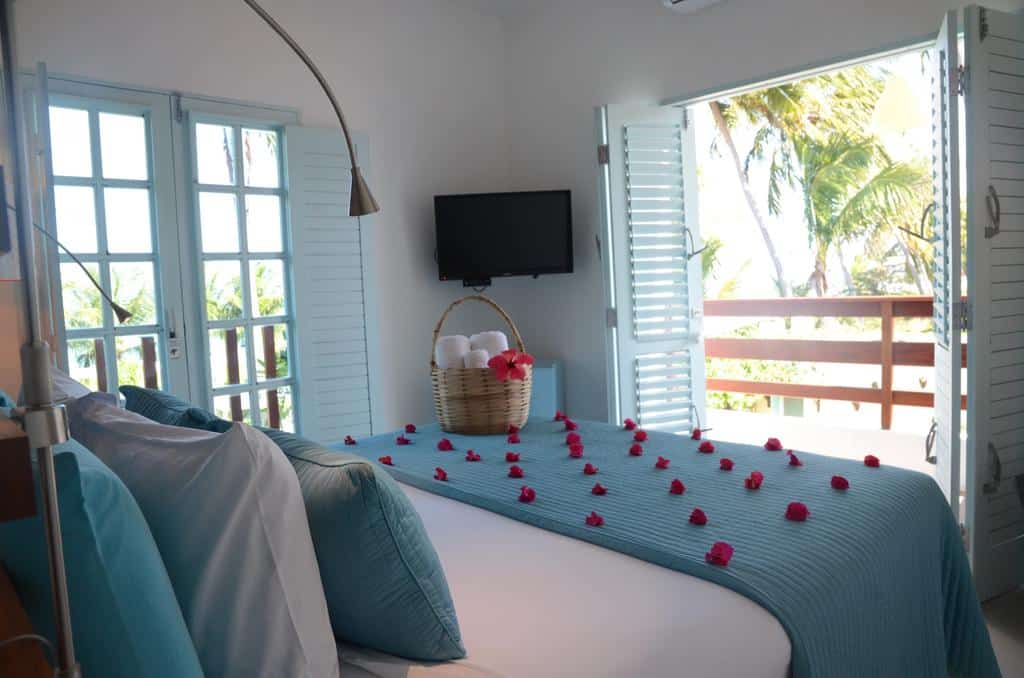 Um quarto com roupa de cama azul, pétalas de rosas na cama, uma cesta com toalhas e persianas abertas mostrando as palmeiras do lado de fora.