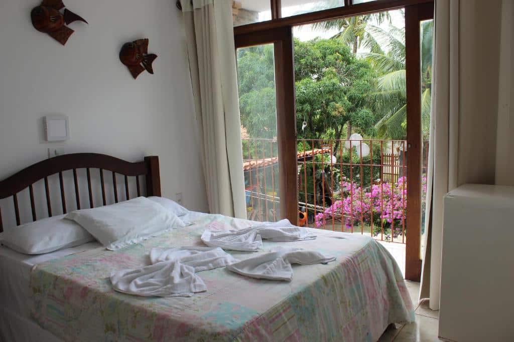 Uma cama com lençóis brancos fica de frente para portas de vidro abertas que dão para uma varanda com flores cor-de-rosa e árvores verdes do lado de fora.