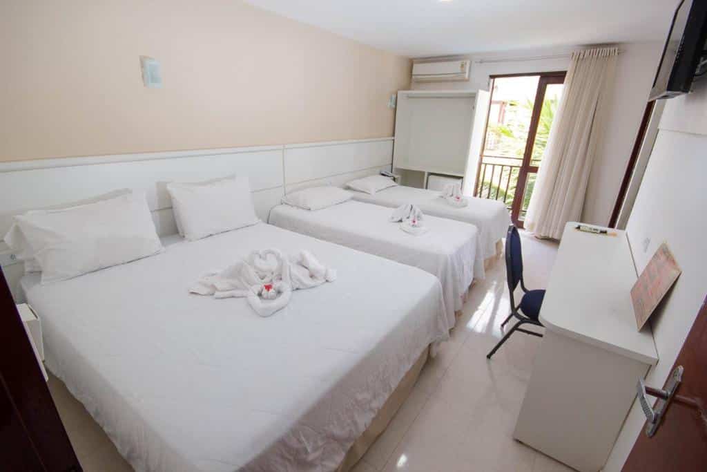 Quarto de hotel com uma cama de casal, duas camas de solteiro, uma escrivaninha, uma cadeira, toalhas dobradas sobre as camas e uma porta de sacada.