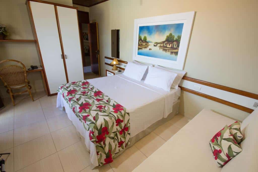 Um quarto de hotel com uma cama de casal, tapete e travesseiro com estampa floral, guarda-roupa, cadeira e obras de arte emolduradas acima da cama.
