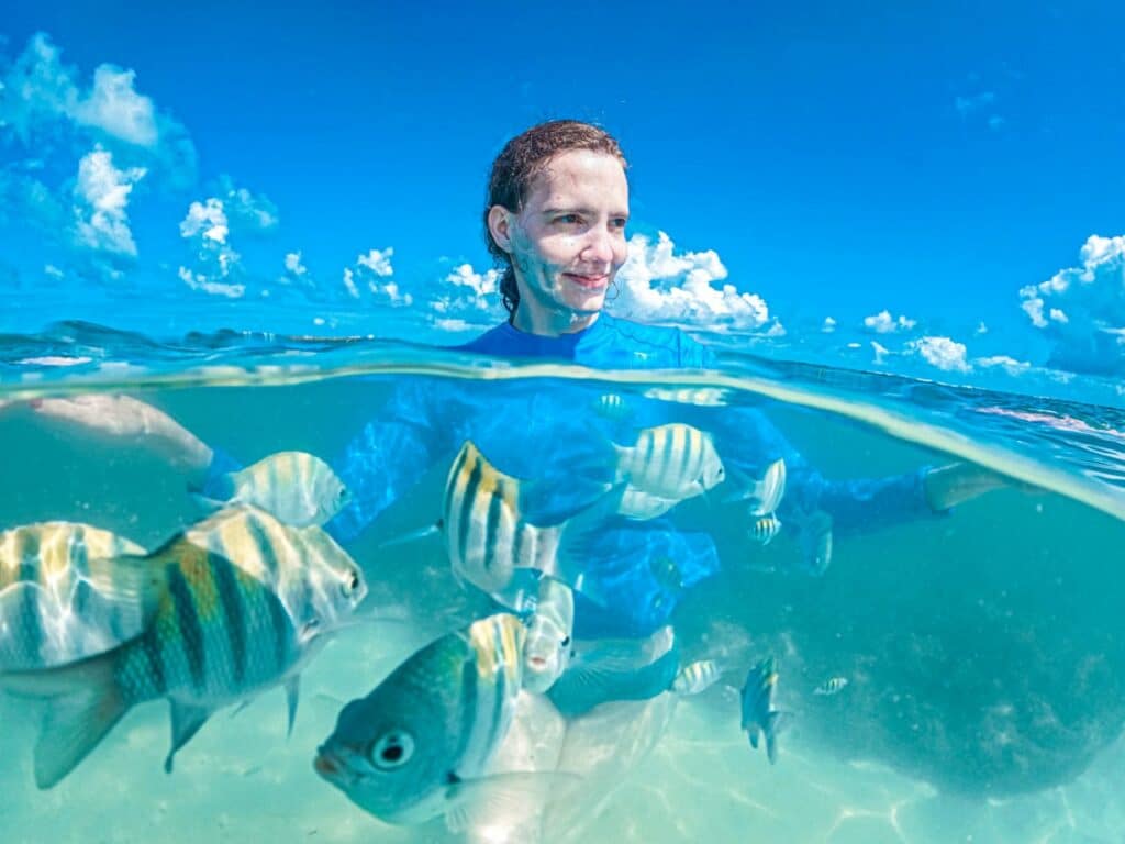 Mulher olhando para o lado, sorrindo, dentro do mar com peixinhos nadando em sua volta. Está de dia e a foto é em uma das piscinas naturais, uma das opções de o que fazer em Maragogi