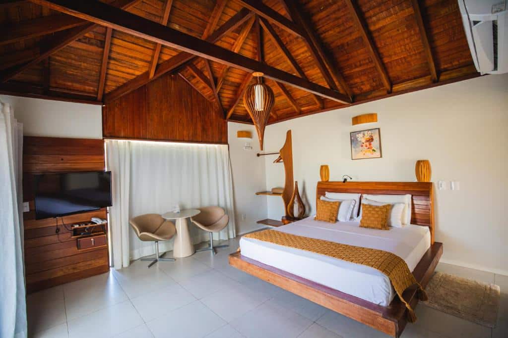 Quarto de hotel moderno com teto de madeira, cama de casal, TV, duas cadeiras, mesa pequena e iluminação aconchegante.