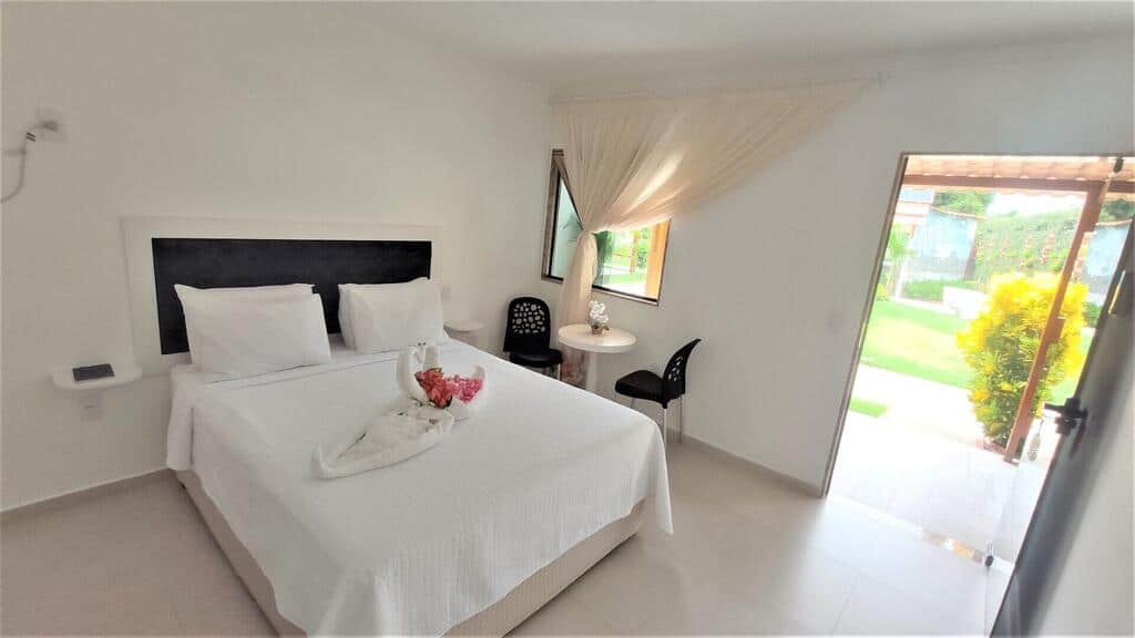 Um quarto bem iluminado com uma cama de casal bem arrumada, uma pequena mesa redonda, duas cadeiras e uma porta aberta que dá para um jardim.