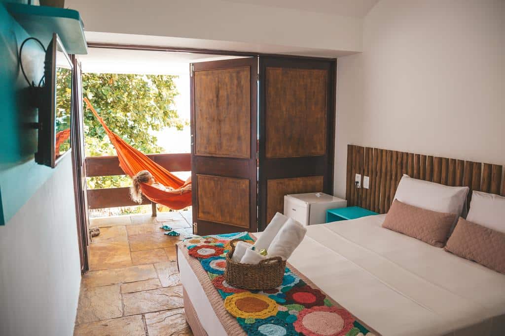 Um quarto com uma cama de casal, cobertor colorido e portas abertas que dão para uma varanda com uma pessoa em uma rede laranja. Esta é uma das pousadas em Maragogi.