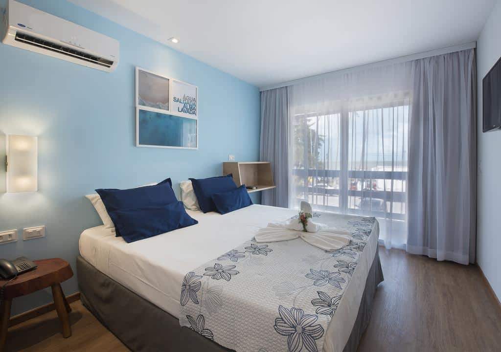 Um quarto de hotel com uma cama de casal, decoração azul e branca, uma escrivaninha, arte na parede e uma varanda com cortinas transparentes.