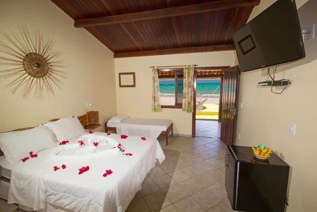 Quarto de hotel com uma cama de casal, cama de solteiro, TV, frigobar e uma porta aberta com vista para a praia do lado de fora.