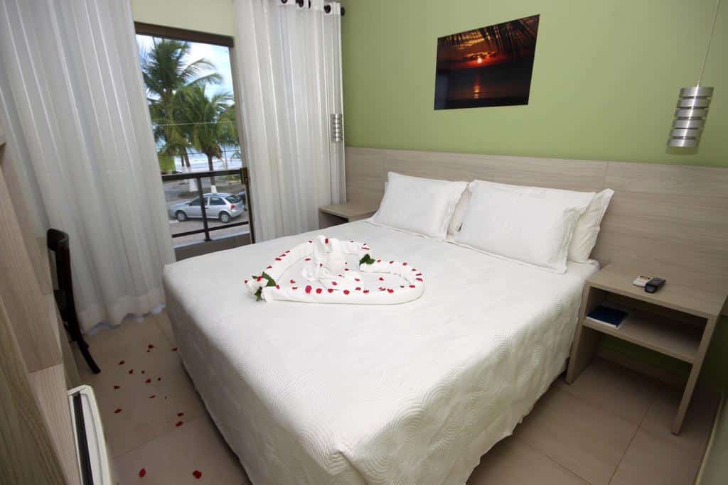Uma cama bem arrumada com lençóis brancos, pétalas de rosa e cisnes de toalha em um quarto de hotel com vista para as palmeiras.