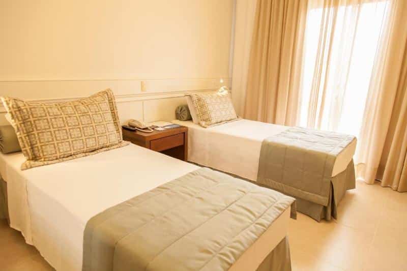 Um quarto de hotel com duas camas de solteiro, uma mesa de cabeceira entre elas e a luz do sol entrando pelas cortinas bege.