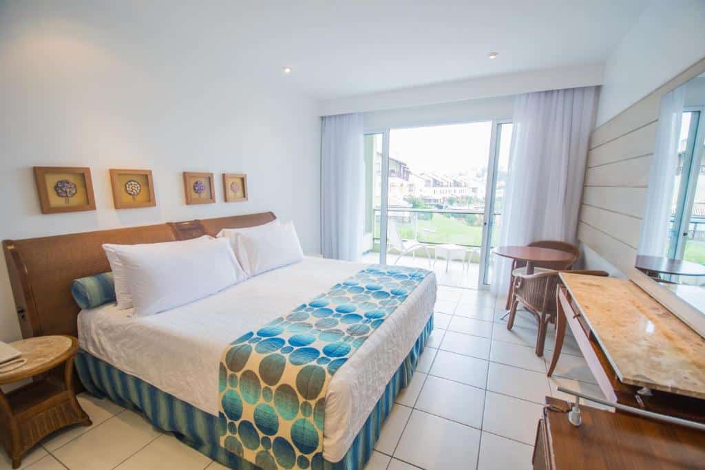 Quarto de hotel bem iluminado com uma cama grande, móveis de madeira, arte na parede e uma varanda com assentos ao ar livre visível através de portas de vidro.