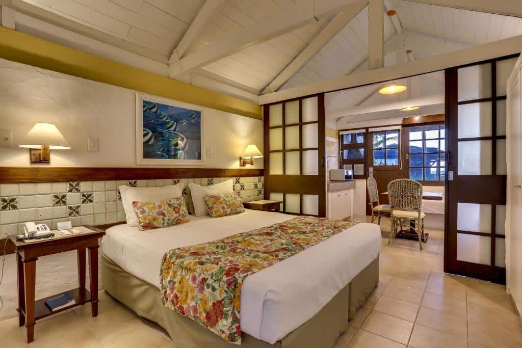 Um quarto de hotel com uma cama de casal, roupa de cama floral, mesas de cabeceira, luminárias e uma escrivaninha perto de portas de correr e uma janela.