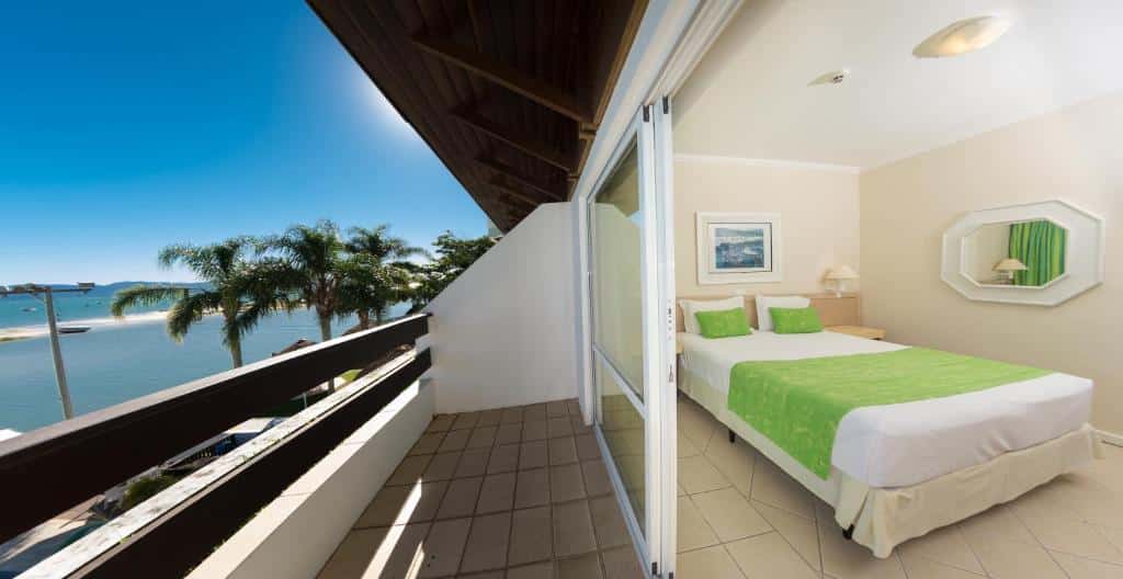 Quarto de hotel com cama de casal, roupa de cama verde e uma varanda com vista para as palmeiras e a água em um dia ensolarado.