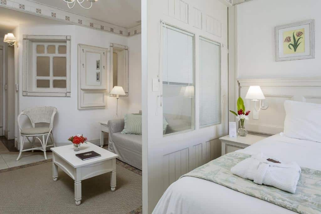 Um quarto de hotel branco e iluminado com uma cama, poltrona, mesa de centro, luminárias e decorações florais.