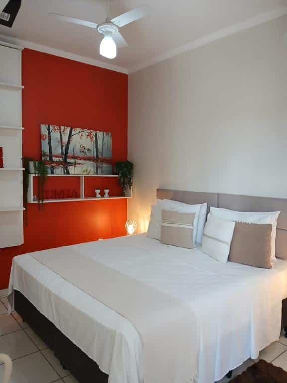 Uma cama bem arrumada com lençóis brancos em um quarto com uma parede vermelha de destaque, arte, prateleiras e travesseiros decorativos, na Hospedagem Quarto Chic centro Cunha, para representar airbnb em Cunha.