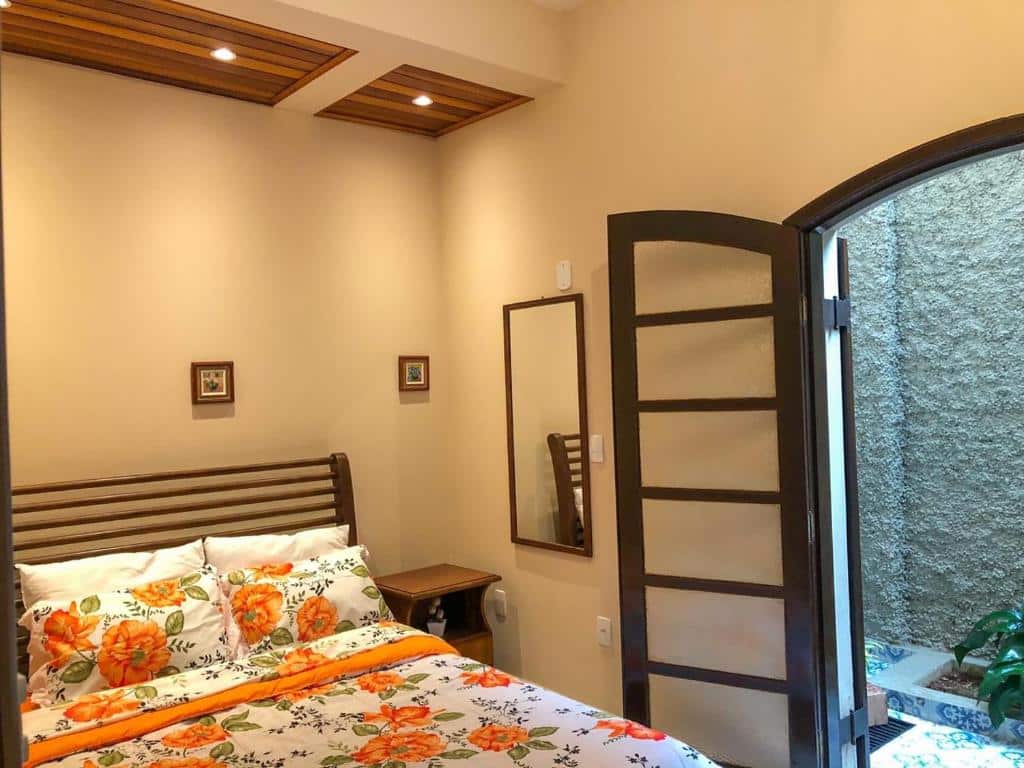 Quarto do Cantinho Cunhense com uma cama de casal com travesseiros e um cobertor florido em cima. Do lado direito da cama tem uma mesinha de madeira, um espelho na parede e uma porta de madeira com vidro que dá acesso a parte externa da casa.