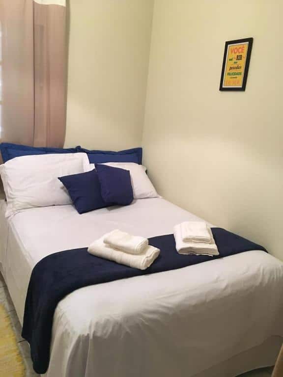Quarto da Hospedagem Aconchego com uma cama de casal com vários travesseiros e almofadas, uma colcha e toalhas em cima. Ao lado na parede tem um quadro e atrás da cama tem uma cortina.