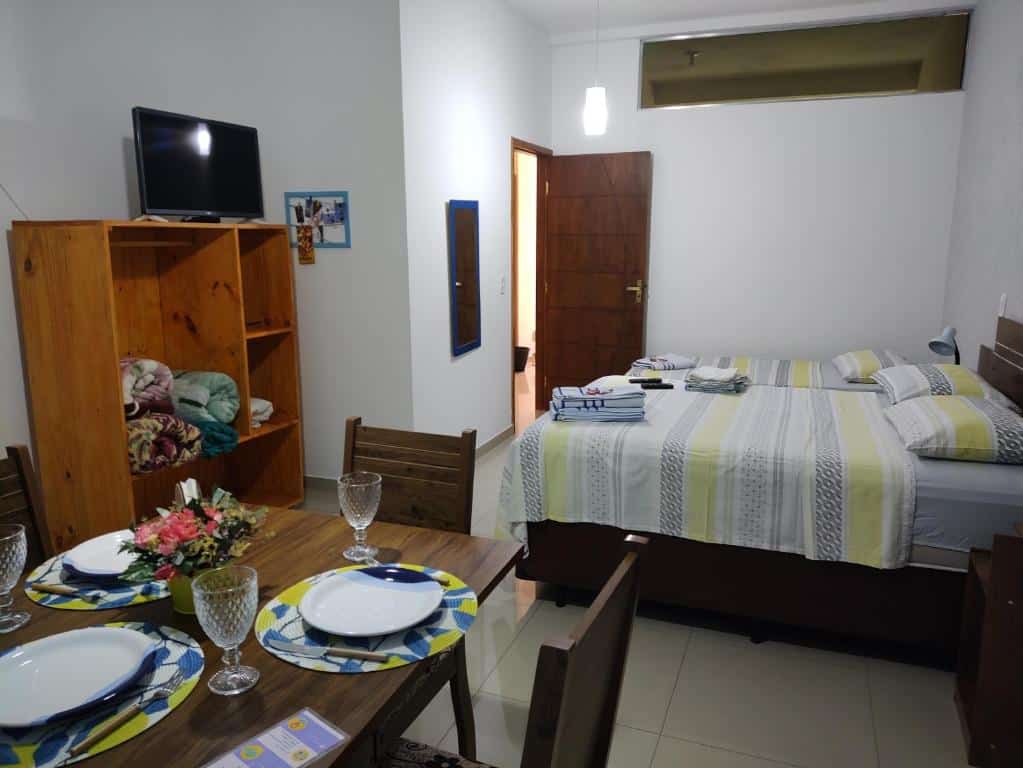 Quarto com 20m² da Hospedagem Elite térreo com uma cama de casal e uma cama de solteiro. No quarto há também uma mesa de madeira quadrada com quatro cadeiras e pratos e copos em cima. Além disso, tem uma ármario aberto com cobertores dentro, uma tv em cima dele e um espelho na parede.