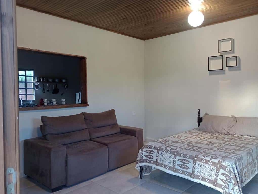 Parte do Sítio Ramos casa amarela que mostra a cama de casal do lado direito da foto e do lado esquerdo um sofá. Atrás do sofá tem um aberto na parede que dá para ver parte da cozinha.