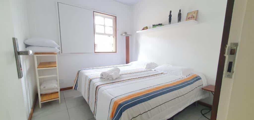 Foto do quarto do Lurgar Apart. Ilustra o post sobre airbnb em Ouro Preto. A cama é box e de casal, e encontra-se no centro. Acima dela há uma prateleira com decorações em cima. Do lado esquerdo dela há uma janela, e em sua frente há uma pequena estante com travesseiros.