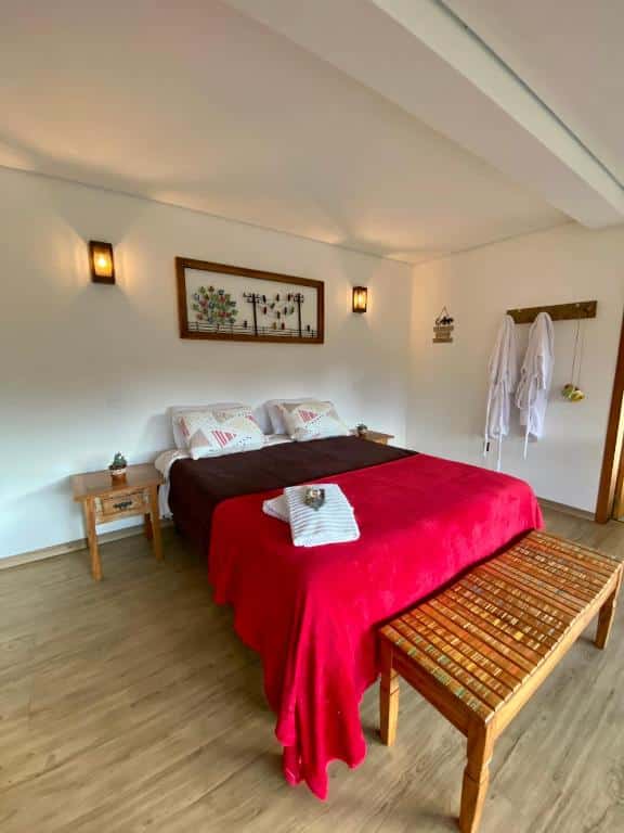 Foto do quarto do CHALÉS SÓ COISAS BOAS. Ilustra o post sobre airbnb em Ouro Preto. A cama está no centro, e ela é box e de casa. Atrás da cama, há um quadro, e do lado direito um gancho que segura dois roupões. Dos dois lados da cama há uma mesa de cabeceira. Encostado no pé da cama está um banco de madeira.