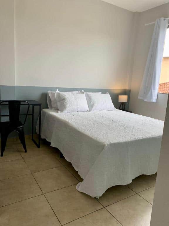 Foto do quarto do Lindo apartamento em frente a UFOP com garagem. Ilustra o post sobre airbnb em Ouro Preto. A cama é box e de casal, e está encostada no meio da parede. Do lado direito dela há uma janela e mesa de cabeceira com abajur. Na esquerda há uma escrivaninha e cadeira.