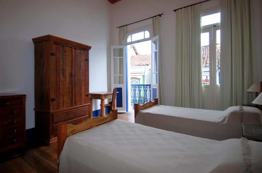 Foto do quarto do Alquimia House. Ilustra o post sobre airbnb em Ouro Preto. Há duas camas de solteiro na direita, e na frente delas está um guarda-roupa, cômoda e mesa com cadeira. No fundo do quarto há duas portas altas com varanda para a rua.