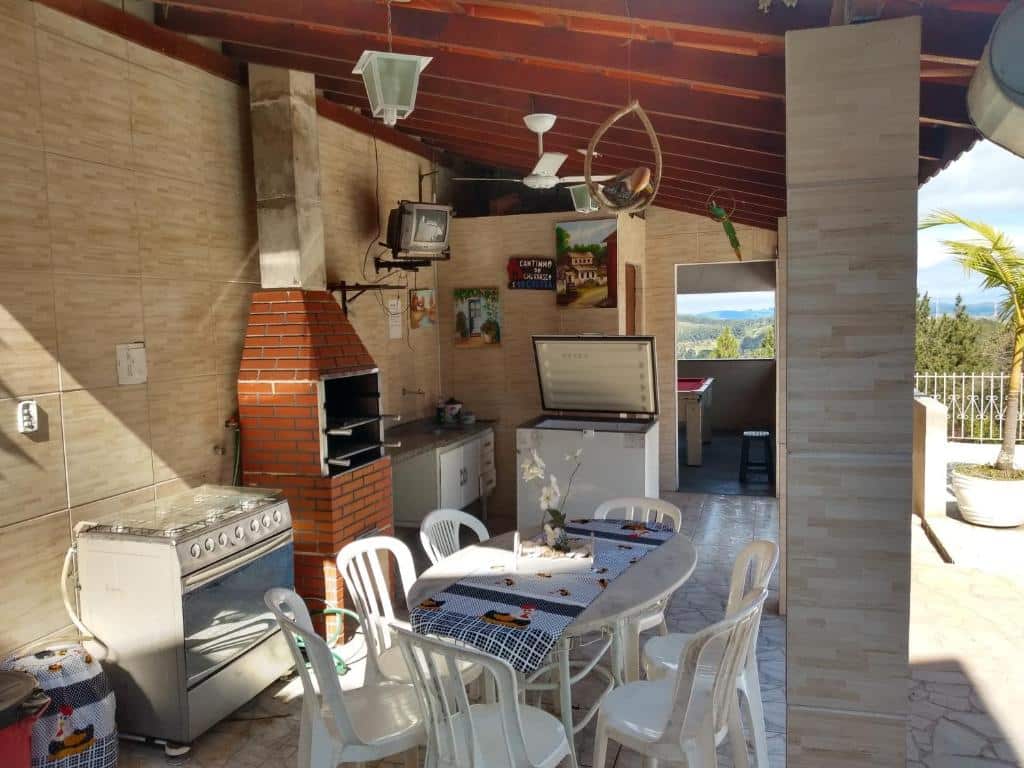 Foto da área de lazer em Chácara Recanto Paraíso. Há uma cozinha ao ar livre com mesa, cadeiras, fogão, churrasqueira, pia e freezer. Também há uma TV no topo de uma parede ao fundo. O rancho é coberto por um telhado. Ilustra o post sobre airbnb em Serra Negra.