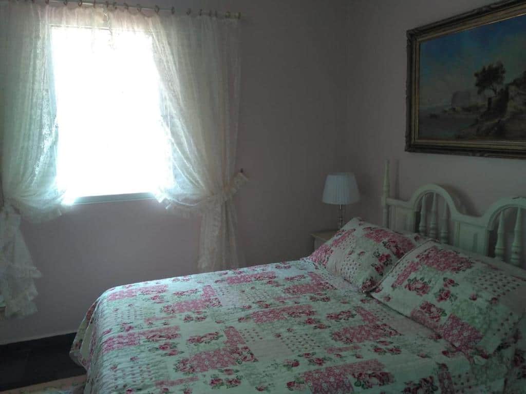 Foto do quarto em Condomínio Vila Paradiso. As paredes são rosa claro. Há uma cama no centro com cabeceira de madeira branca, e ao seu lado na direita há um gaveteiro branco de madeira. Há um quadro atrás da cama, decorando a parede. Ilustra o post sobre airbnb em Serra Negra.