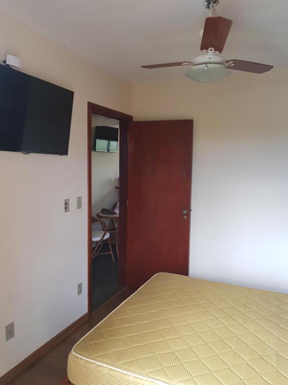 Foto do quarto em Piemonte Flat. Há uma cama de casal no canto inferior direito, vemos o colchão de relance. De frente para a cama há uma TV fixa na parede. No teto há um ventilador. porta também está de frente com a cama. Ilustra o post sobre airbnb em Serra Negra.