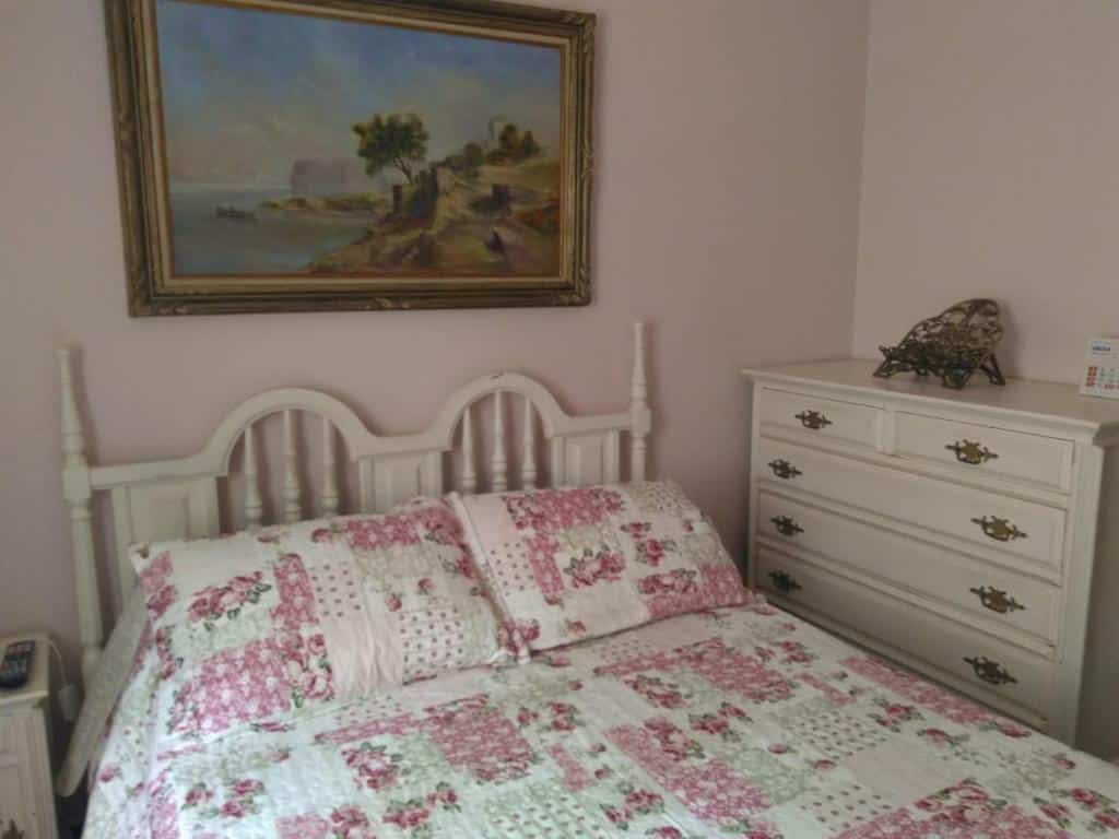 Foto do quarto em Condomínio Vila Paradiso. As paredes são rosa claro. Há uma cama no centro com cabeceira de madeira branca, e ao seu lado na direita há um gaveteiro branco de madeira. Há um quadro atrás da cama, decorando a parede. Ilustra o post sobre airbnb em Serra Negra.
