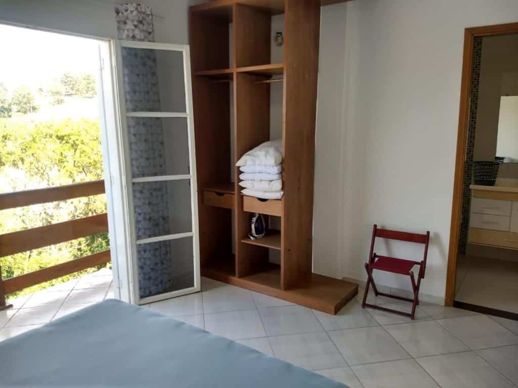 Foto do quarto em Casa com linda Vista no Centro. A cama está no canto inferior esquerdo vemos parte do pé do colchão. De frente há uma estante de madeira com almofadas, e de seu lado esquerdo há uma varanda. Na direita há uma porta para o banheiro. Ilustra o post sobre airbnb em Serra Negra.