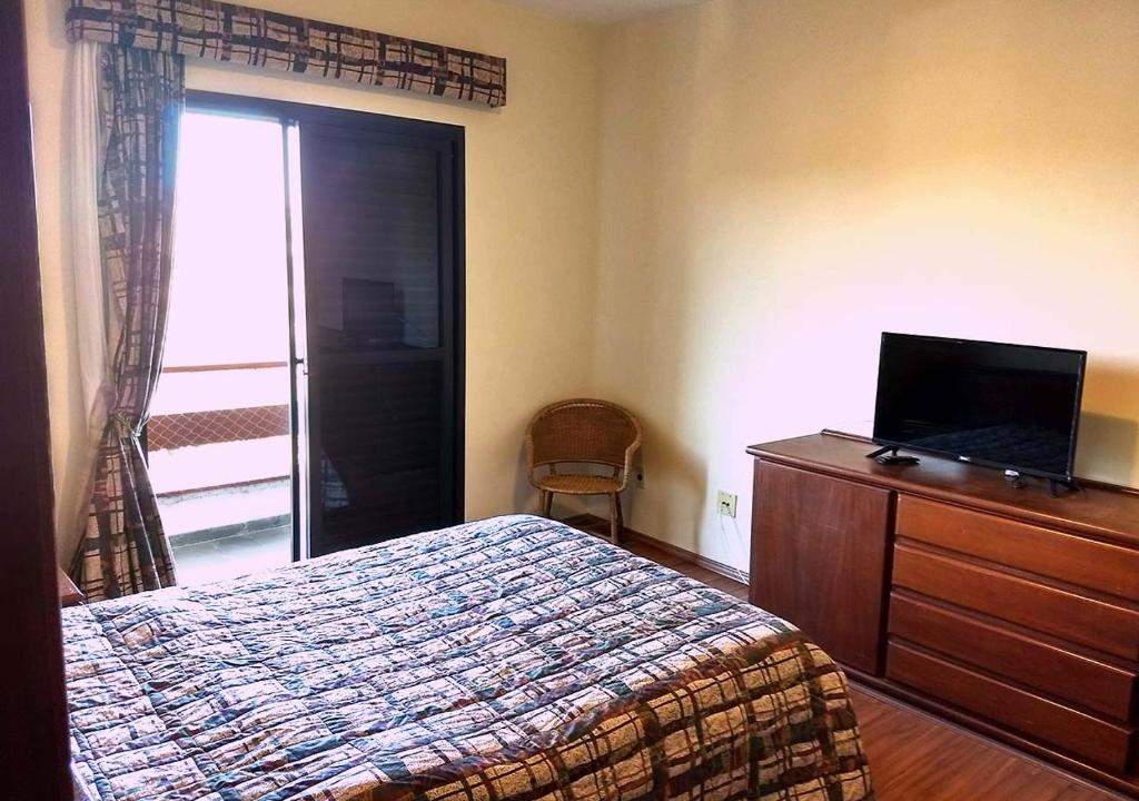 Foto do quarto em Piemonte Flat Serra Negra. A cama está no meio, vemos ela de lado. Na frente dela há uma estante de madeira com uma pequena TV em cima. Do outro lado do quarto há uma porta com varanda. Ilustra o post sobre airbnb em Serra Negra.