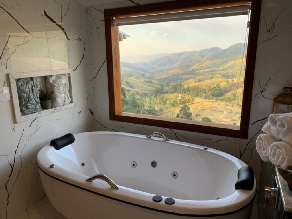 Banheiro do Hotel Pousada Shangri-la com uma banheira de hidromassagem e atrás uma janela com vista para as montanhas.