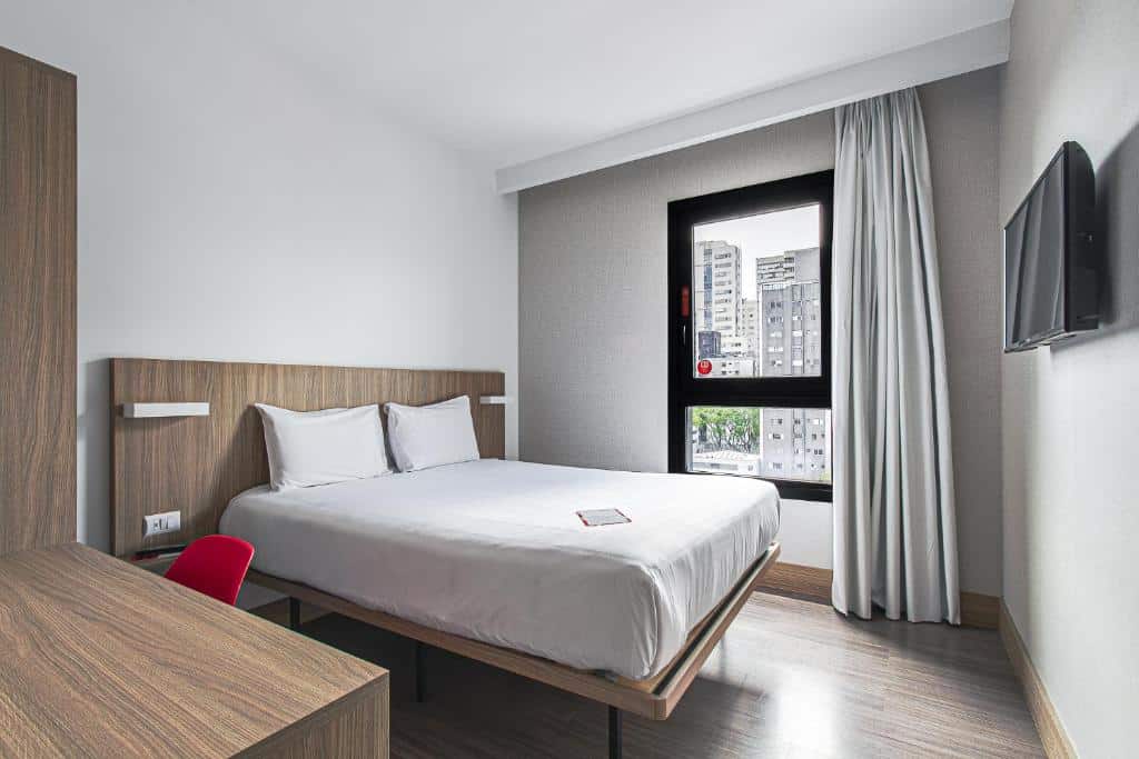 Quarto do Hotel Moov Curitiba. Uma cama de casal do lado esquerdo e uma mesa de trabalho. Do lado direito uma televisão e no fundo a janela do quarto. Foto para ilustrar post sobre hotéis em Curitiba.