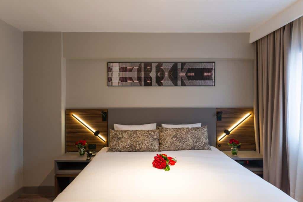 Quarto do Mercure Curitiba Batel. Uma cama de casal no meio com uma flor em cima. De cada lado uma cômoda e uma luminária. No lado direito a janela do quarto.