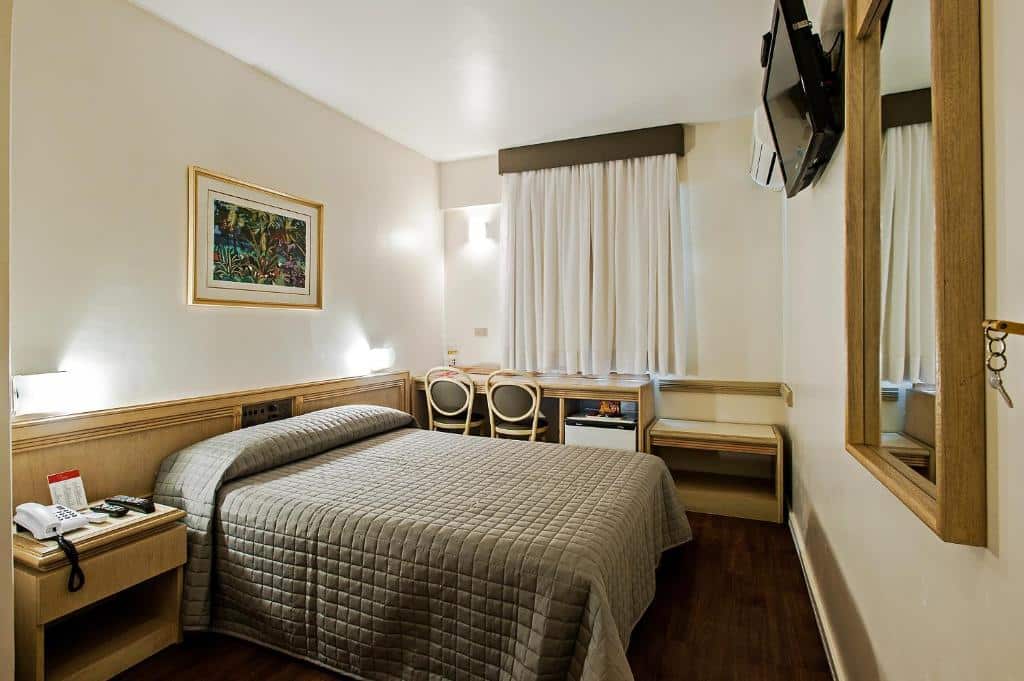 Quarto do Trevi Hotel e Business. Uma cama de casal do lado esquerdo, com um espelho de frente, do lado do espelho uma televisão e o ar-condicionado. No fundo, uma mesa de trabalho e um frigobar. Em cima, a janela do quarto.