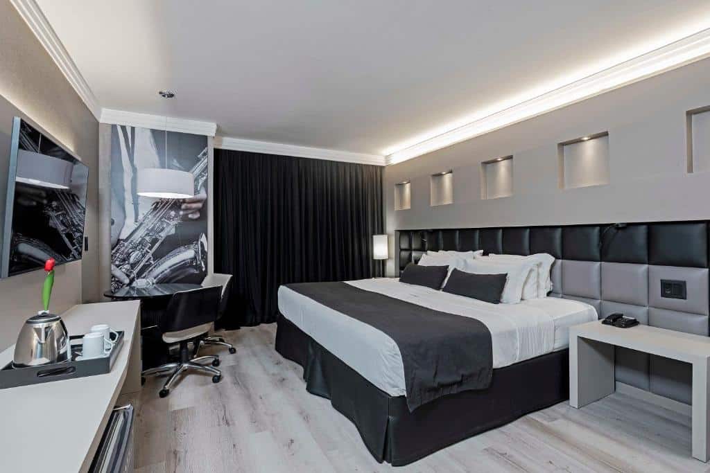 Quarto do FULL JAZZ. Uma cama de casal do lado direito, do lado esquerdo uma raque, um frigobar, uma bandeja com xícaras e em cima uma televisão. No fundo, uma mesa com duas cadeiras, luminária e a janela do quarto com cortina fechada. Foto para ilustrar post sobre hotéis em Curitiba.
