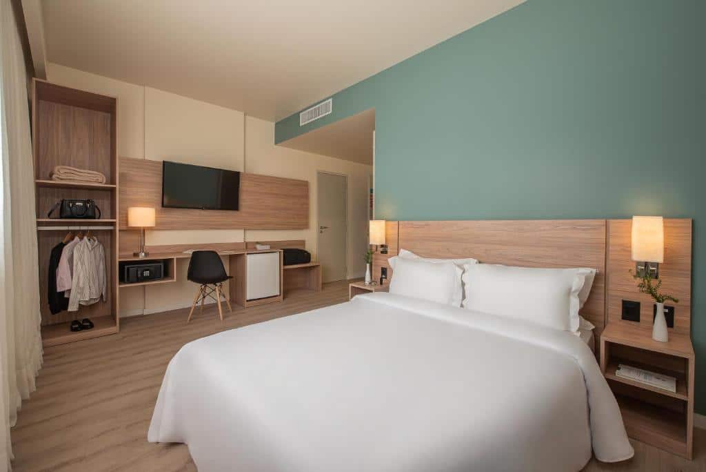 Quarto do Intercity Curitiba Batel. Uma cama de casal do lado direito, com uma luminária e estante de cada lado. No fundo, um armário, um cofre, uma mesa de trbalho com frigobar, em cima um televisão. Foto para ilustrar post sobre hotéis em Curitiba.