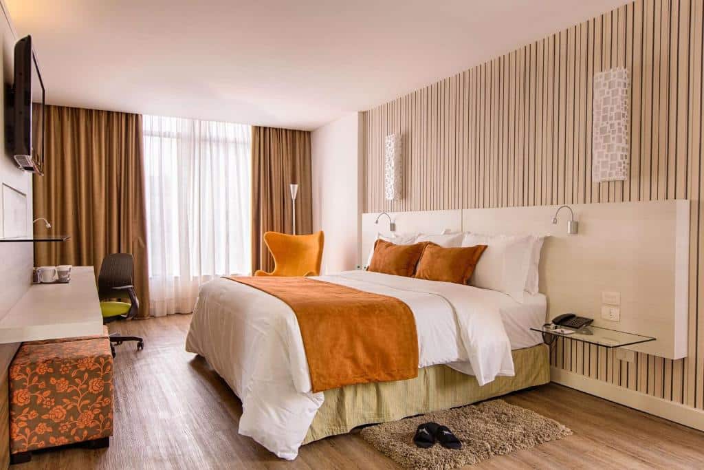 Quarto do Radisson Hotel Curitiba. Uma cama de casal do lado direito com uma estante e uma luminária de cada lado. No fundo uma poltrona e a janela do quarto. Do lado direito uma televisão, uma estante, e a mesa de trabalho. Foto para ilustrar post sobre hotéis em Curitiba.