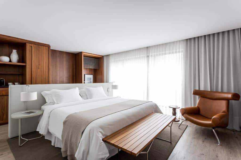 Quarto do Nomaa Hotel. Uma cama de casal do lado esquerdo com um banco na frente e de frente uma poltrona. De cada lado da cama uma mesinha e uma luminária. No fundo, atrás da cama, um guarda-roupa. Foto para ilustrar post sobre hotéis em Curitiba.