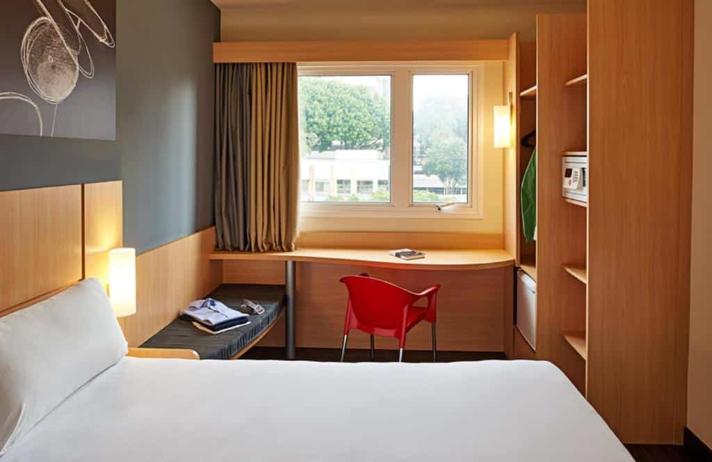 Quarto do ibis Curitiba Batel. Uma cama de casal do lado esquerdo, e um banco no fundo. Do lado direito um guarda-roupa com cofre e frigobar. No fundo, uma mesa de trabalho e em cima a janela do quarto. Foto para ilustrar post sobre hotéis em Curitiba.